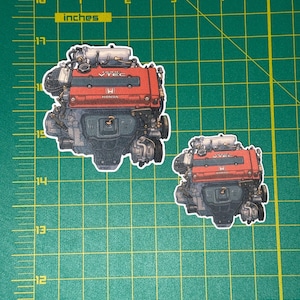 May include: Two die-cut stickers of a Honda engine, one large and one small. The engine is red and gray with the Honda logo and "VTEC" text visible. The stickers are on a green cutting mat with inch measurements.