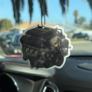 May include: An air freshener featuring a detailed illustration of a car engine. The design is in shades of gray and black, with a black string for hanging. The air freshener is hanging in a car.