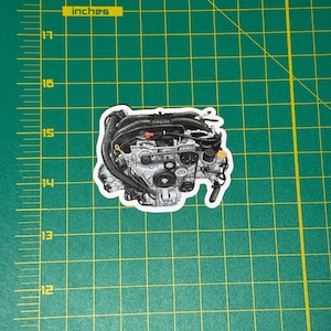 May include: A black and white sticker of a car engine. The sticker is on a green surface with yellow inch markings. The engine has various components, including belts, hoses, and a complex mechanical design. The sticker is approximately 4 inches wide.