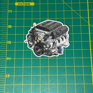 May include: A sticker featuring a detailed illustration of a car engine. The engine is depicted in shades of gray, black, and silver, showcasing various components like belts, pulleys, and the engine block. The sticker is on a green cutting mat with inch measurements.