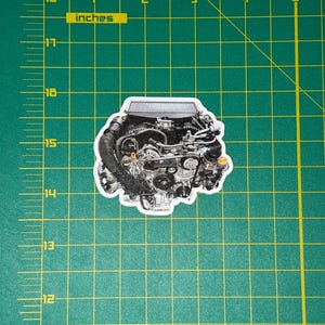 May include: A sticker featuring a detailed illustration of a car engine. The engine is depicted in shades of gray and black, with various mechanical components visible. The sticker is placed on a green grid background with inch measurements.