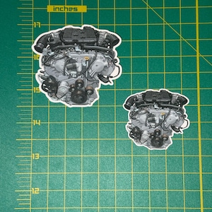 May include: Two die-cut stickers featuring detailed illustrations of a car engine. The stickers are in grayscale, showcasing the engine's intricate components. The stickers are placed on a green grid background with the word "inches" visible.