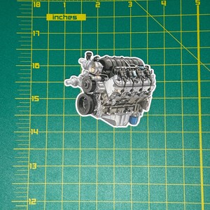 May include: A sticker featuring a detailed illustration of an engine, showcasing various mechanical components. The engine is primarily silver with black and blue accents. The sticker is approximately 5 inches in length.