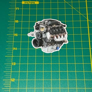 May include: A sticker featuring a detailed illustration of a car engine. The engine is primarily silver and black, with red accents on the exhaust headers and a blue component. The sticker is placed on a green cutting mat with inch measurements.