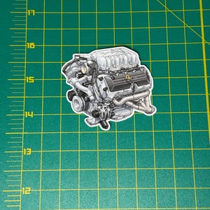 May include: A sticker featuring a detailed illustration of a car engine. The engine is primarily gray with black accents and the text "POWER BY FORD". The sticker is placed on a green surface with inch measurements.