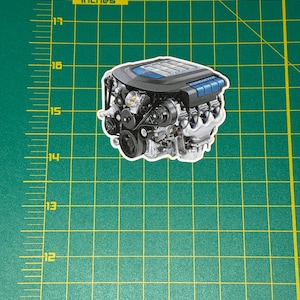 May include: A sticker of a detailed car engine, featuring black, silver, and blue components. The engine is approximately 3 inches wide and 4 inches long. The sticker is on a green cutting mat with inch markings.