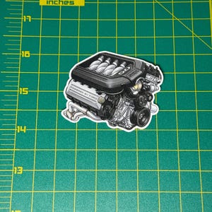 May include: A sticker of a detailed car engine, predominantly black and silver, with the text "POWERED BY FORD". The engine is depicted with various components, including exhaust pipes and belts. The sticker is on a green cutting mat with inch markings.