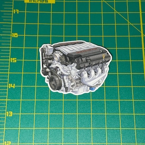 May include: A sticker featuring a detailed illustration of a car engine. The engine is primarily silver and black, with intricate mechanical components visible. The sticker is on a green grid background with inch measurements.