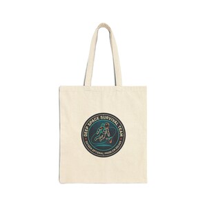 Pode incluir: Saco tote creme com um emblema circular com a ilustração de um astronauta. O emblema diz "DEEP SPACE SURVIVAL TEAM" e "OXYGEN OPTIONAL PROBLEM SOLVER" em verde-azulado e vermelho.