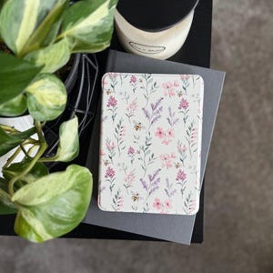 May include: A rectangular, floral-patterned case featuring a design of pink and purple flowers, green leaves, and bees on a white background. The case is resting on a gray book. A candle and a potted plant are in the background.