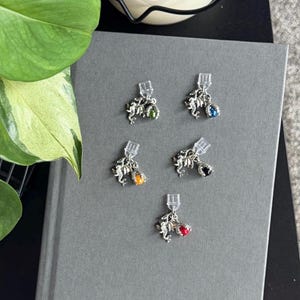 May include: Five silver-toned metal charms, each featuring a detailed dragon figure clutching a small, round, colored bead. The beads are green, blue, orange, black, and red. Each charm is attached to a clear plastic clip for easy attachment.