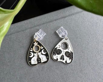 Pode incluir: Um par de brincos em forma de tabuleiro Ouija em preto e branco. Um brinco apresenta dois gatos brancos, luas e estrelas. O outro brinco tem as palavras "YES" e "NO" com detalhes florais. Os brincos têm costas de plástico transparente.