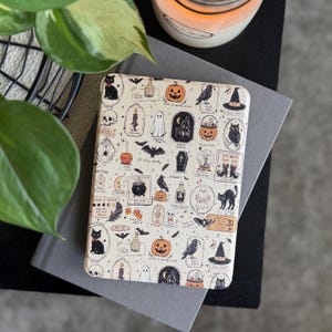 May include: A rectangular e-reader case featuring a cream-colored background with a repeating pattern of Halloween-themed illustrations including black cats, pumpkins, ghosts, bats, skulls, ravens, witch hats, cauldrons, and various small text labels.
