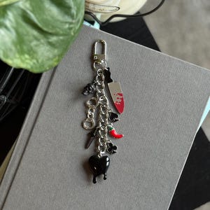 May include: A silver keychain with multiple charms, including a skull, a knife with red accents, a chili pepper, and a black heart. The keychain is attached to a silver clasp.