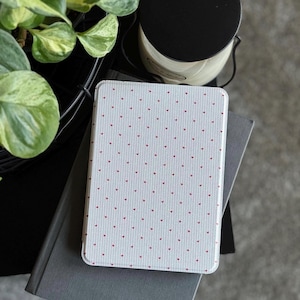 May include: A white tablet case with a pattern of small red hearts. The case is resting on top of a gray book. A black candle and a green plant are in the background.