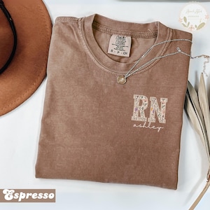 May include: Espresso-colored t-shirt with the text "RN ashley" in a floral design. The shirt has a crew neck and short sleeves. A silver necklace with a pendant is on the shirt.