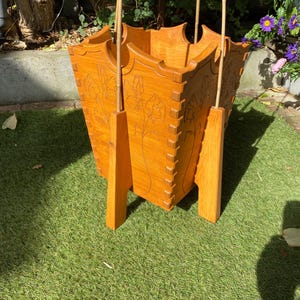 May include: A light brown wooden umbrella stand. The stand has a square shape with carved floral designs and is supported by four wooden legs. The stand is on a green surface.