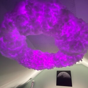 May include: A cloud-shaped light fixture with a vibrant purple glow. The fixture is mounted on a white ceiling. A black and white moon art print hangs on the wall below the light.
