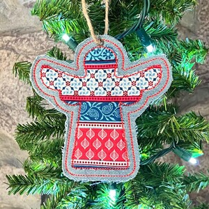 Puede incluir: Un adorno de ángel de tela hecho a mano con un diseño estampado en rojo, azul y blanco. El adorno está colgado de un árbol de Navidad artificial verde.