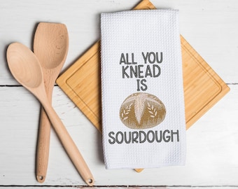 May include: A white waffle-weave kitchen towel featuring an embroidered golden-brown sourdough loaf graphic and the black text "ALL YOU KNEAD IS SOURDOUGH" positioned above and below the bread illustration, accompanied by two light-colored wooden spoons.