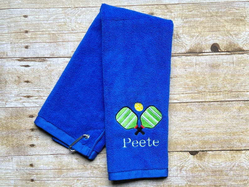 Op de afbeelding: Een blauwe handdoek met een pickleball-ontwerp geborduurd in groen, geel en zwart. Het woord "Peete" is eronder geborduurd. De handdoek heeft een lus en ligt op een houten oppervlak.