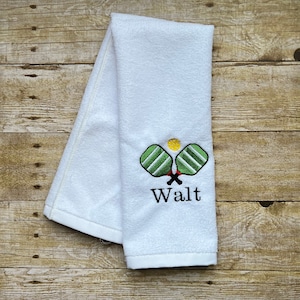 Op de afbeelding: Witte handdoek met geborduurde pickleball rackets en een gele bal. De rackets zijn groen en wit, met rode accenten. De naam "Walt" is eronder geborduurd.