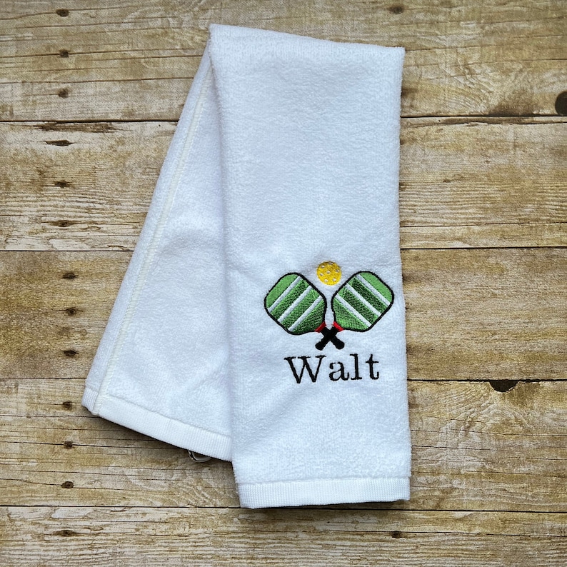 Op de afbeelding: Witte handdoek met geborduurde pickleball rackets en een gele bal. De rackets zijn groen en wit, met rode accenten. De naam "Walt" is eronder geborduurd.