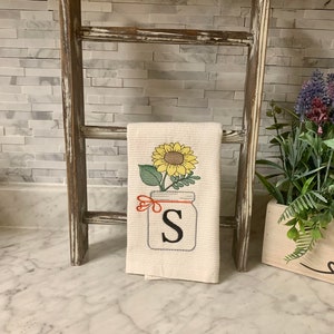 Puede incluir: Una toalla de cocina blanca con un girasol y un tarro de cristal con la letra "S" bordada. La toalla está colgada en una escalera de madera rústica.