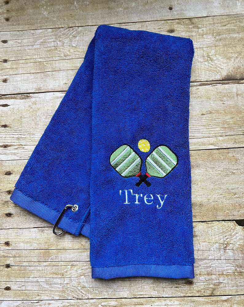 May include: Royal blue towel with an embroidered pickleball design. The design includes two green and white pickleball paddles, a yellow ball, and the name "Trey" in white script. A silver carabiner clip is attached.