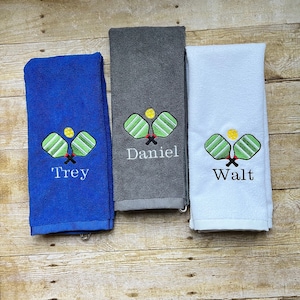 May include: Three folded towels in blue, gray, and white. Each towel features an embroidered pickleball design with a yellow ball and green paddles. The blue towel has "Trey" embroidered, the gray towel has "Daniel", and the white towel has "Walt".
