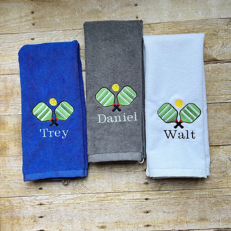 May include: Three folded towels in blue, gray, and white. Each towel features an embroidered pickleball design with a yellow ball and green paddles. The blue towel has "Trey" embroidered, the gray towel has "Daniel", and the white towel has "Walt".