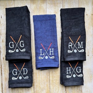 Peut inclure: Serviettes de golf bleues marine et noires, brodées d'initiales et de motifs de clubs de golf. Les serviettes présentent des clubs de golf croisés, une balle de golf et des initiales personnalisées en fil blanc. Les serviettes sont pliées.