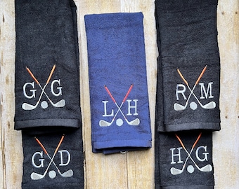 May include: Golf towels in navy blue and black, embroidered with initials and golf club designs. The towels feature crossed golf clubs, a golf ball, and personalized initials in white thread. The towels are folded and displayed on a wooden surface.