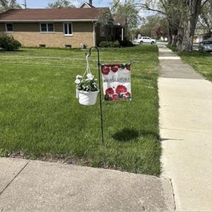 May include: A black metal garden stake holds a white hanging flower pot and a rectangular flag. The flag displays the word "welcome" and red floral designs. The scene is set on a green lawn with a house in the background.