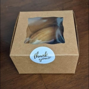 May include: A small, square, brown cardboard box with a clear window, containing baked goods. A blue and white sticker on the front reads "thank you" in cursive. The box is closed and ready for gifting.