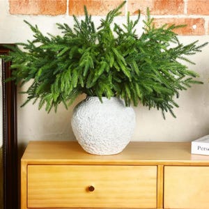 Könnte beinhalten: Eine weiße, strukturierte Vase gefüllt mit leuchtend grünen künstlichen Tannenzweigen. Die Vase steht auf einer hellen Holzkommode, mit einer Backsteinmauer im Hintergrund. Das Arrangement bringt ein Naturelement in den Raum.