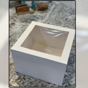 May include: A white, square bakery box with a clear window on top. The box has a scalloped edge around the window and is designed to hold baked goods. The box is sitting on a speckled countertop.
