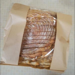 May include: Sliced loaf of bread in a clear plastic bag, wrapped in a brown paper bag. The bread is light brown with a textured crust. The slices are visible through the plastic.