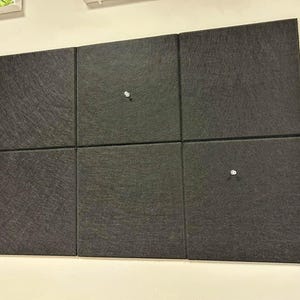 May include: Six dark gray square acoustic panels mounted on a white wall. The panels are arranged in a rectangular grid and secured with silver tacks. The panels have a textured surface.