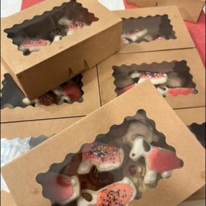 May include: Several kraft paper boxes with scalloped window cutouts, filled with decorated cookies. The cookies are in various shapes, including dog and heart shapes, with pink, white, and brown icing and sprinkles.