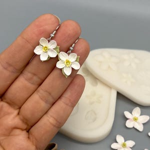 Könnte beinhalten: Ein Paar weiße Blumenohrringe mit gelber Mitte, begleitet von zwei weißen Silikonformen mit Blumen- und Blattmotiven, mehreren losen weißen Blumenverzierungen und drei braunen, blütenblattförmigen Ausstechformen.