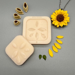 Può includere: Uno stampo in silicone quadrato color beige con motivo a petali, quattro piccoli tagliapasta a forma di petalo, un ciondolo a forma di girasole su catenina argentata e diversi petali in argilla gialli e verdi disposti in linea curva.