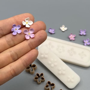 Könnte beinhalten: Kleine vierblättrige Blumenformen in Weiß und verschiedenen Violetttönen, zusammen mit zwei weißen Silikonformen mit blumenförmigen Vertiefungen und vier braunen Kunststoff-Ausstechformen für Bastel- und Knetprojekte.