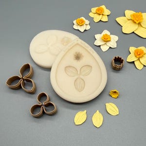 Könnte beinhalten: Zwei beige Silikonformen mit geprägten Blumenmustern, zwei braune dreilappige Metallausstecher, ein kleiner brauner runder Ausstecher sowie mehrere gelbe und cremefarbene Narzissenblütenblätter und zusammengesetzte Blüten.