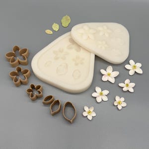 Könnte beinhalten: Zwei cremeweiße Silikonformen mit eingeprägten Blumen- und Blattformen, dazu mehrere braune Kunststoff-Ausstechformen für Blumen und Blätter sowie eine Ansammlung kleiner, weißer fünfblättriger Blüten mit gelben Mitten daneben.