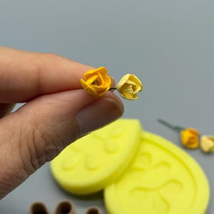 Może przedstawiać: Dwa miniaturowe sztuczne kwiaty w odcieniach jasnego i bladego żółtego, w towarzystwie dwóch żółtych silikonowych form z kwiatowymi odciskami oraz dwóch brązowych plastikowych wykrawaczy w kształcie trójlistnych płatków.