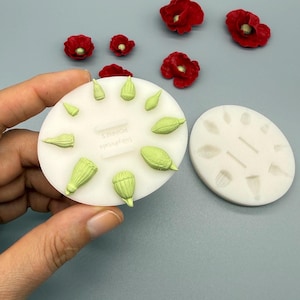 Könnte beinhalten: Zwei weiße Silikonformen zum Basteln, die jeweils mehrere Vertiefungen zur Herstellung von Mohnkapseln aufweisen. Eine Form ist mit hellgrünen Modelliermasse-Formen gefüllt, daneben liegen mehrere kleine rote Mohnblumen-Dekorationen.