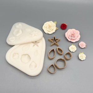 Può includere: Una collezione di materiali per creare decorazioni floreali. Include due stampi in silicone bianchi con impronte di fiori e foglie, diversi taglierini in metallo marrone di varie forme e fiori di carta fatti a mano nei toni del rosa, rosso e crema.