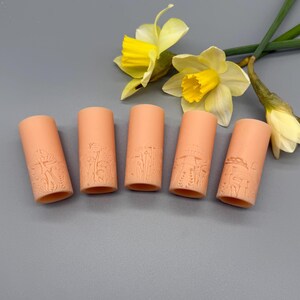 May include: Five cylindrical peach-colored clay beads featuring embossed mushroom and botanical designs arranged in a horizontal row, accompanied by two yellow daffodil flowers with green stems positioned in the upper right corner.