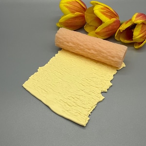 Può includere: Un mattarello color pesca chiaro con una superficie strutturata poggia su un foglio di argilla gialla. L'argilla ha un motivo strutturato simile. Tre tulipani con petali gialli e rossi sono sullo sfondo.
